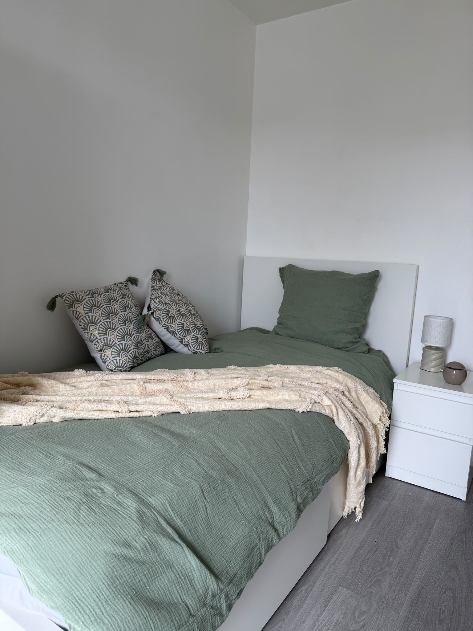 Een eenpersoonsbed met een groene plaid en twee kussens met een patroon, een witte nachtkastje met een lamp, en een beige deken.