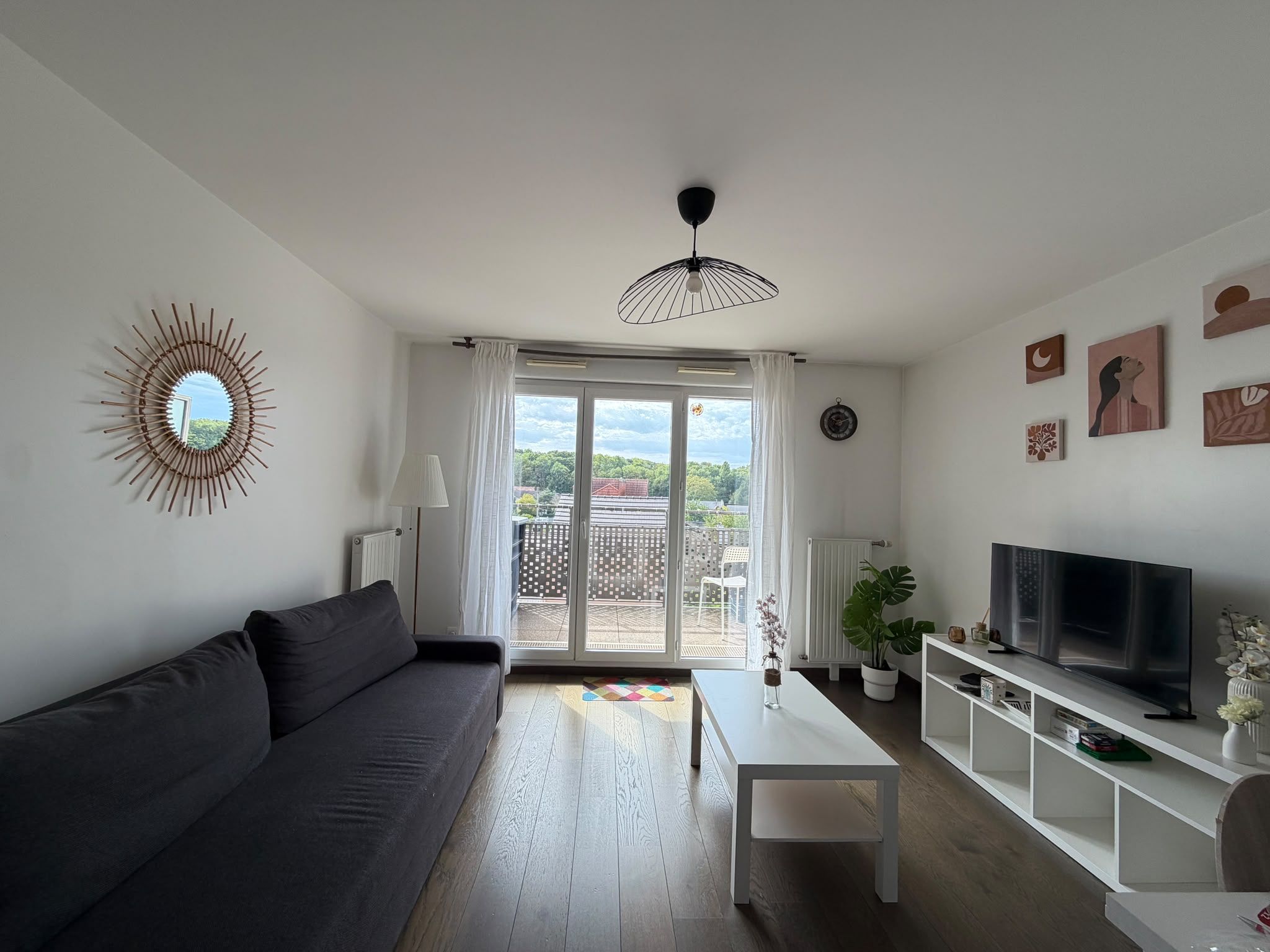 A modern living room with a sofa, a coffee table, a TV on a stand, and a balcony with a view.