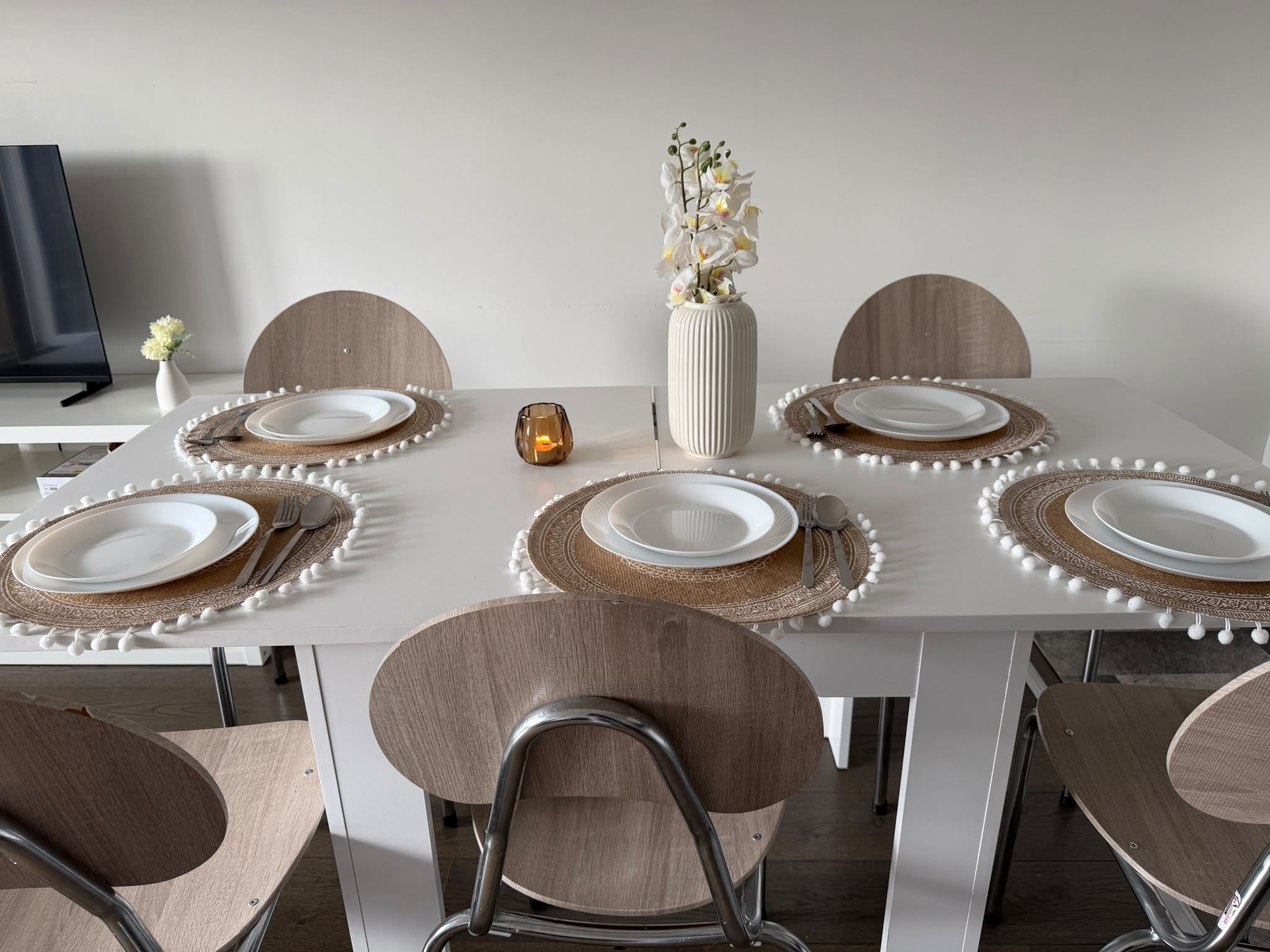 A dining table set with white plates, brown placemats, and a vase of white flowers.