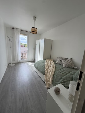 A bedroom with a bed, a bedside table, a hanging light fixture, a coat rack, and a window with curtains. - L'Escale Parisienne: a perfect 2-bedroom apartment in charming Tremblay (Tremblay-en-France)