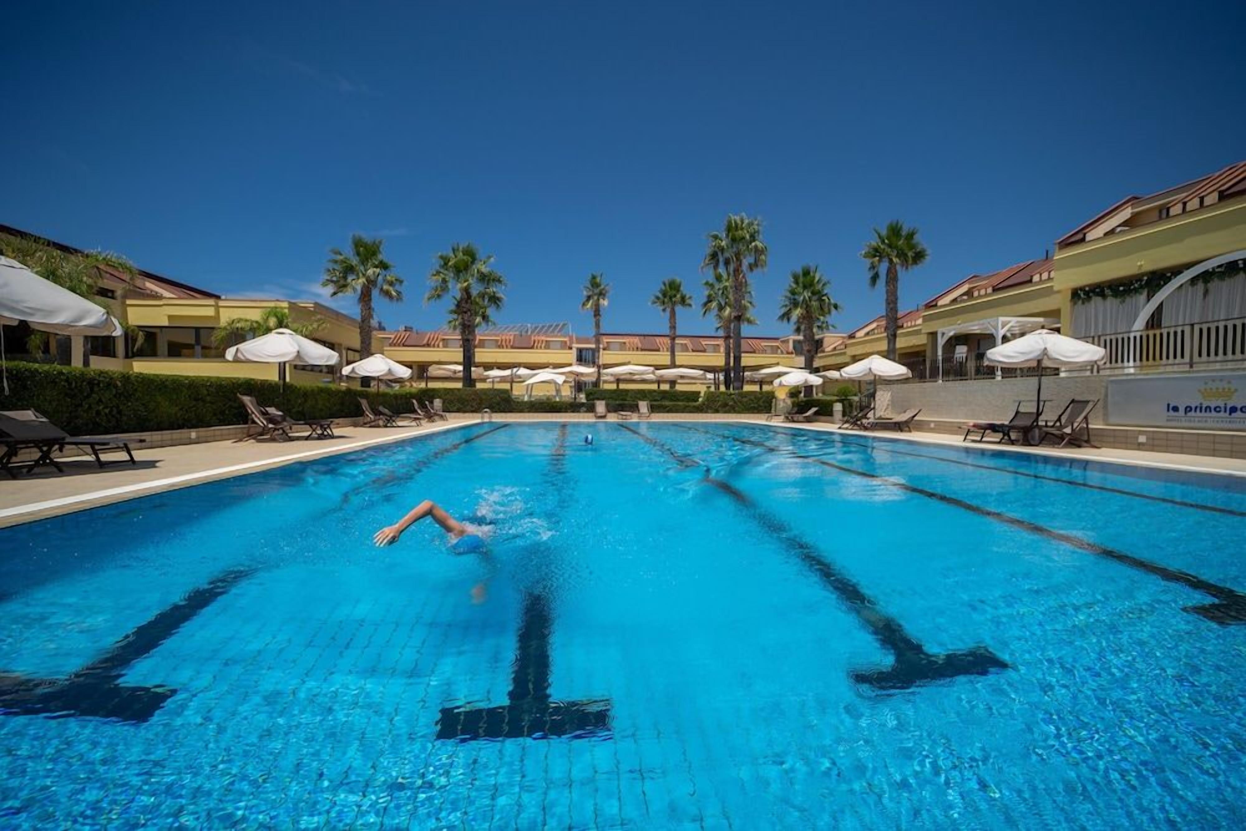 Une piscine entourée de palmiers et des bâtiments de l’hôtel, avec des chaises longues et des parasols.