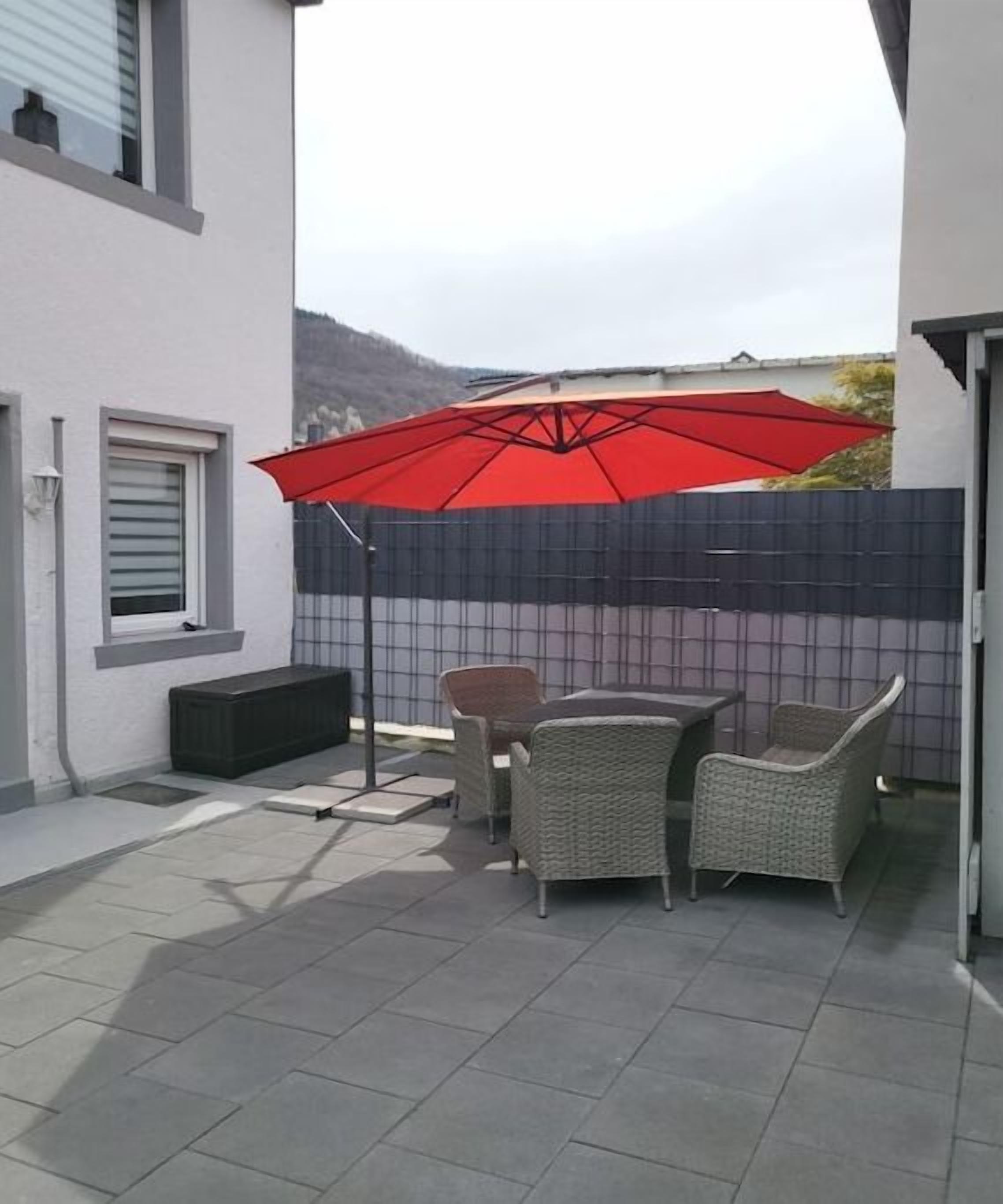 Een terras buiten met een rode parasol, rieten meubels en uitzicht op de omgeving.