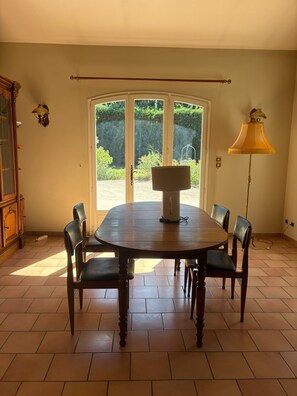 Een eethoek met een ronde tafel, zes stoelen, een lamp en uitzicht door een glazen deur.