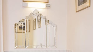 Un baño con lavamanos pedestal, un espejo con marco decorativo y una mesita de noche con lámpara y un libro.