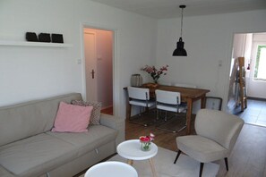 A living room with a sofa, a chair, a small table with flowers, a dining area with chairs, and a hanging light fixture. - Holiday apartment with 2 bedrooms (Dishoek)
