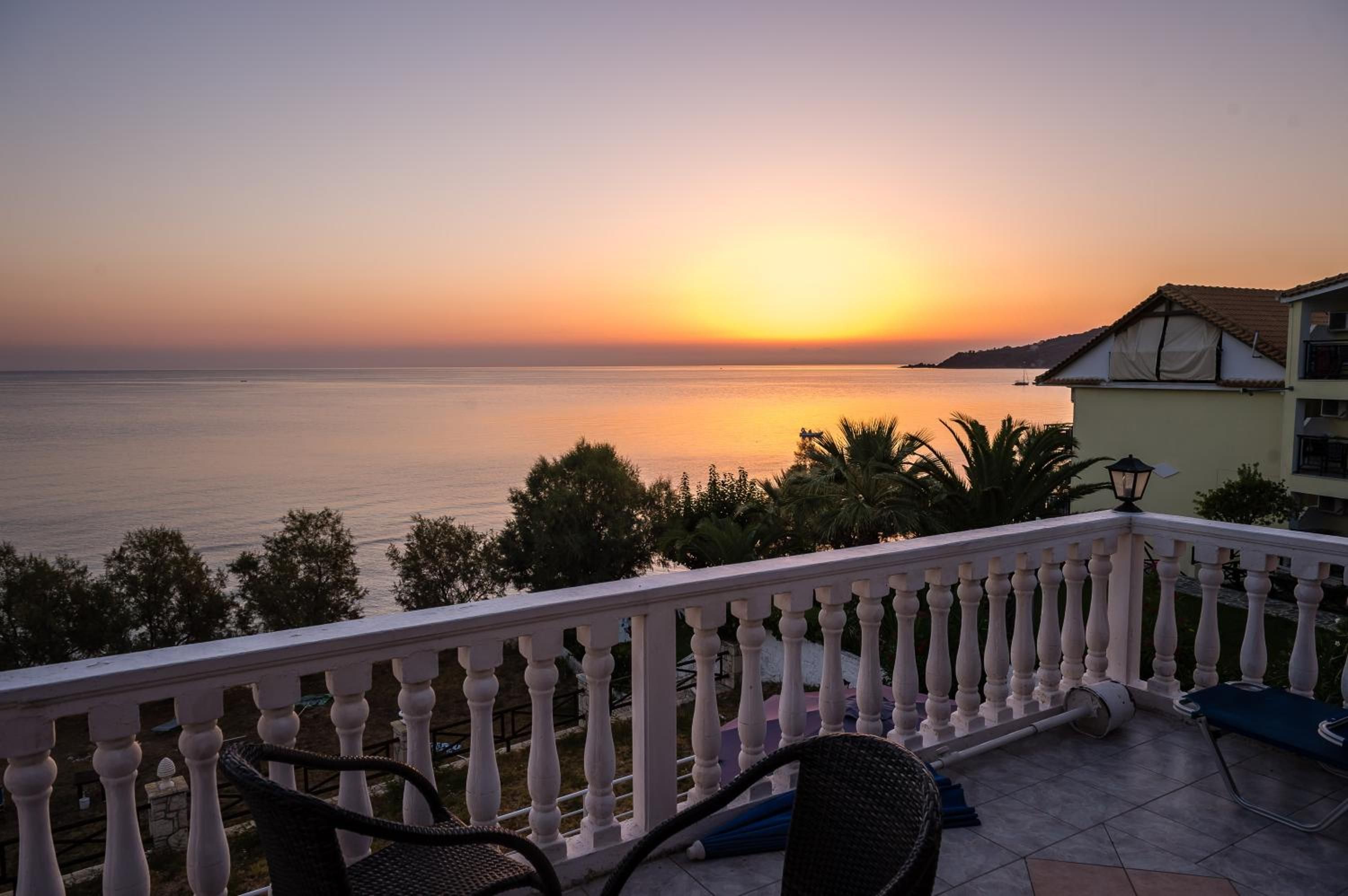 A balcony with a view of the sea during sunset, featuring a railing, chairs, and a building in the background.