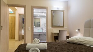 Habitación de hotel con cama, escritorio, silla, espejo y baño visible a través de una puerta abierta.