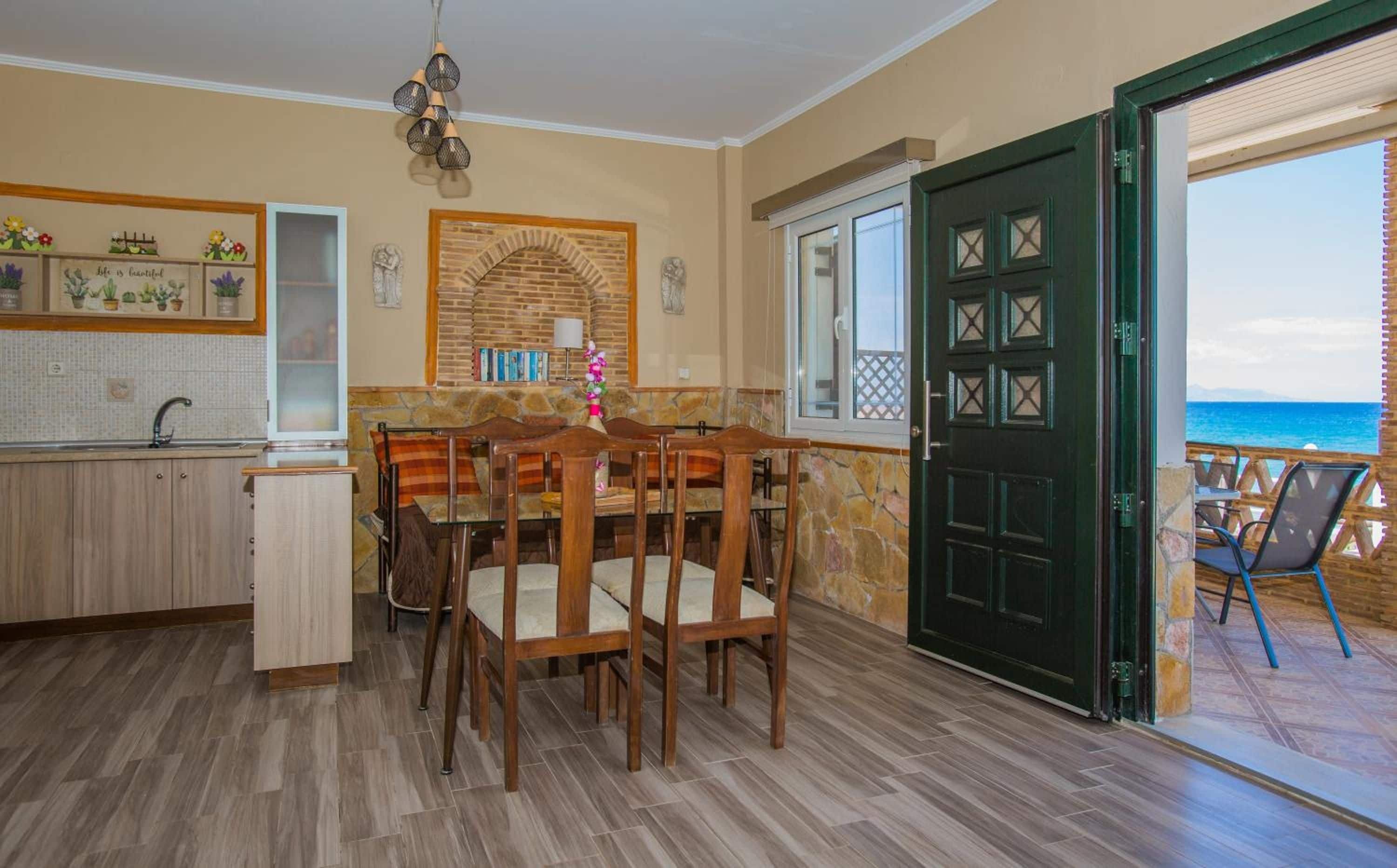 A dining area with a table and chairs, a kitchen with a sink and cabinets, and a door leading to a balcony with a view of the sea.