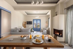 A modern living room with a dining table set for breakfast, a fireplace, and a view through a blue door. - Ambeli Villa (Santorini)