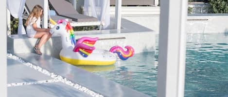 A pool area with a unicorn float, a child sitting on a lounge chair, and a poolside structure.