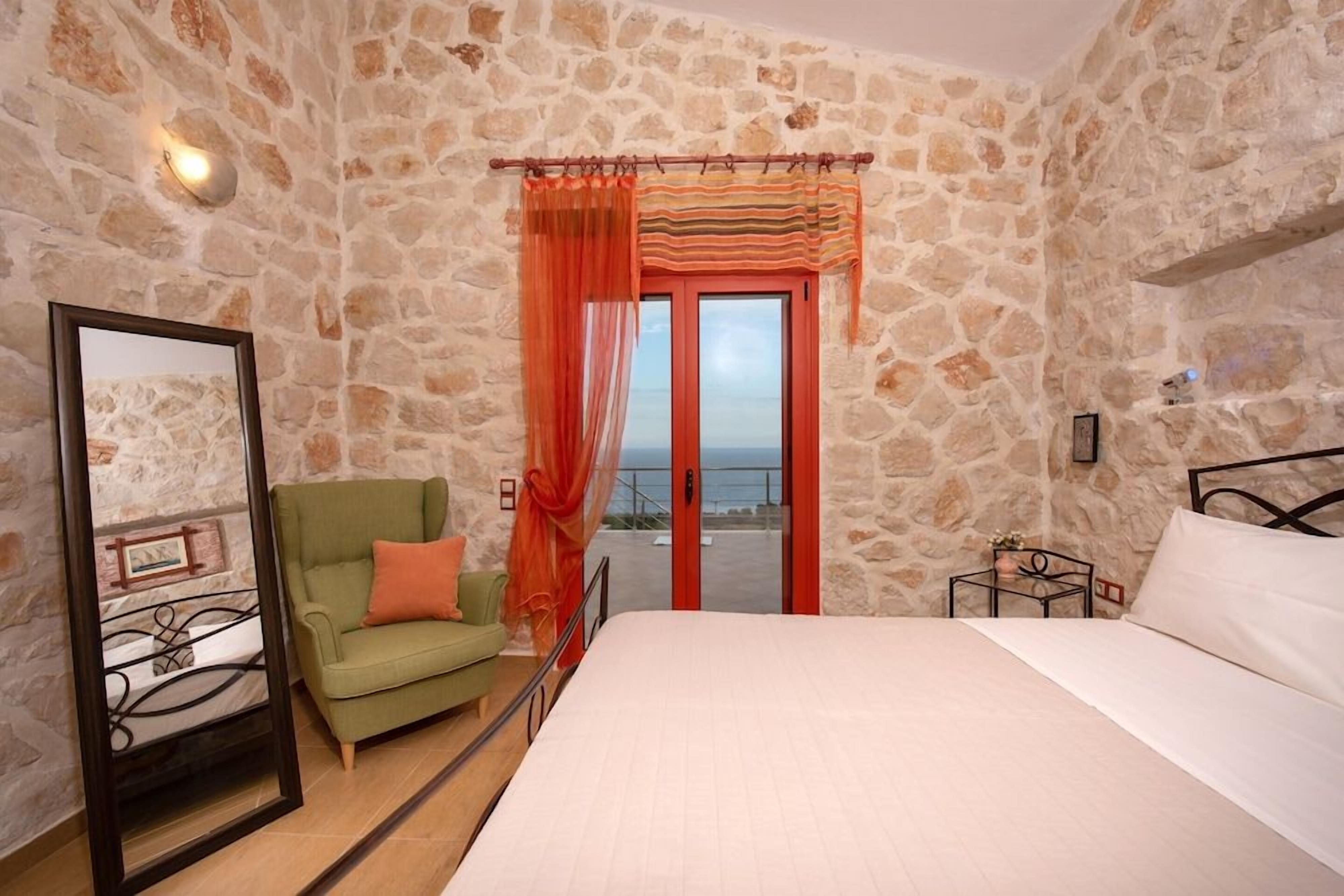 A bedroom with a stone wall, a bed, a green armchair, a mirror, and a view through a door.