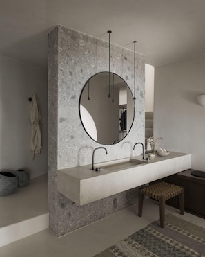 A bathroom with a large, round mirror, a modern sink, and a bench. - Vora Private Villas (Santorini)