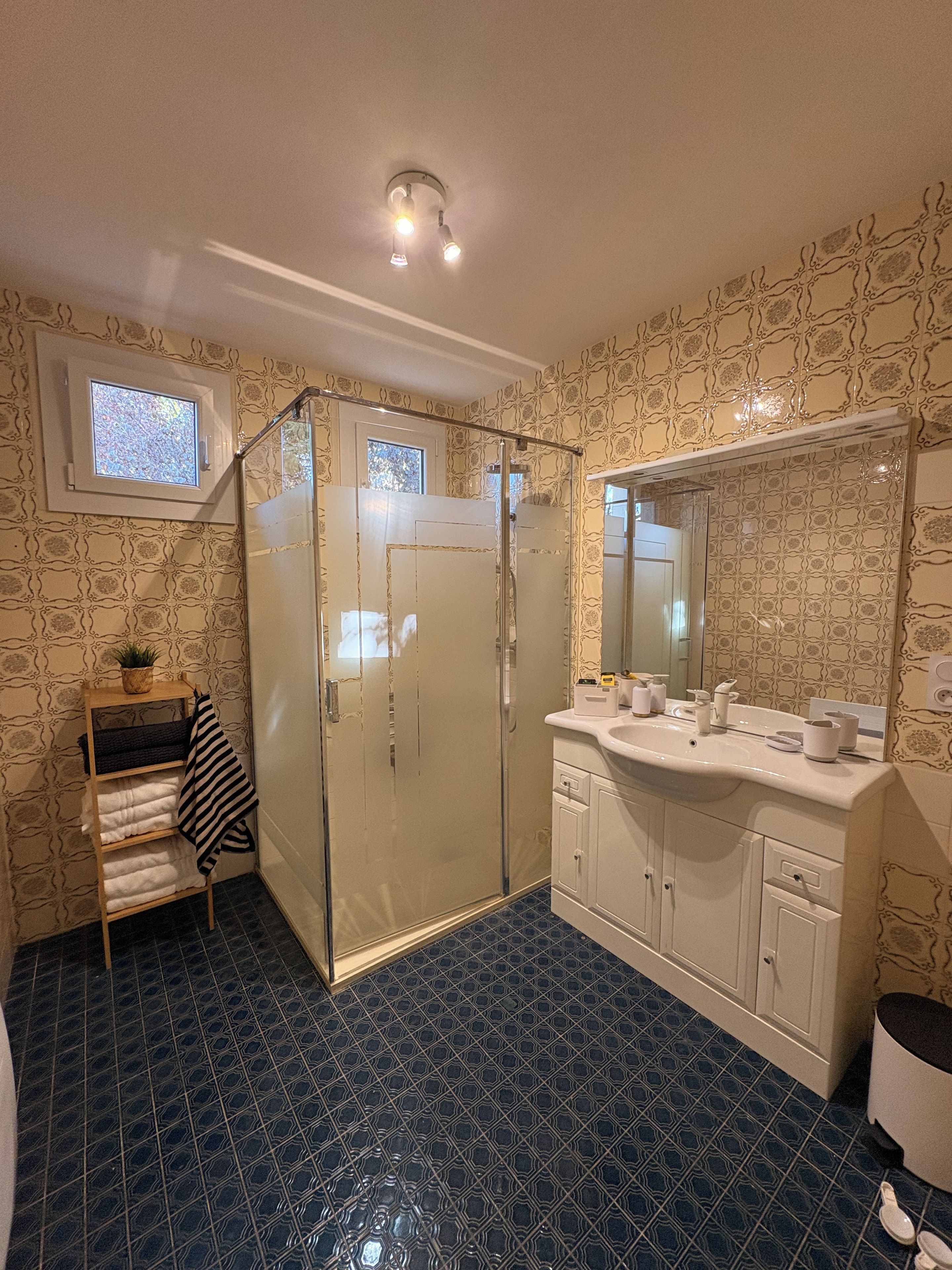 A bathroom with a glass shower enclosure, a white sink with a mirror, patterned wallpaper, and a small window.