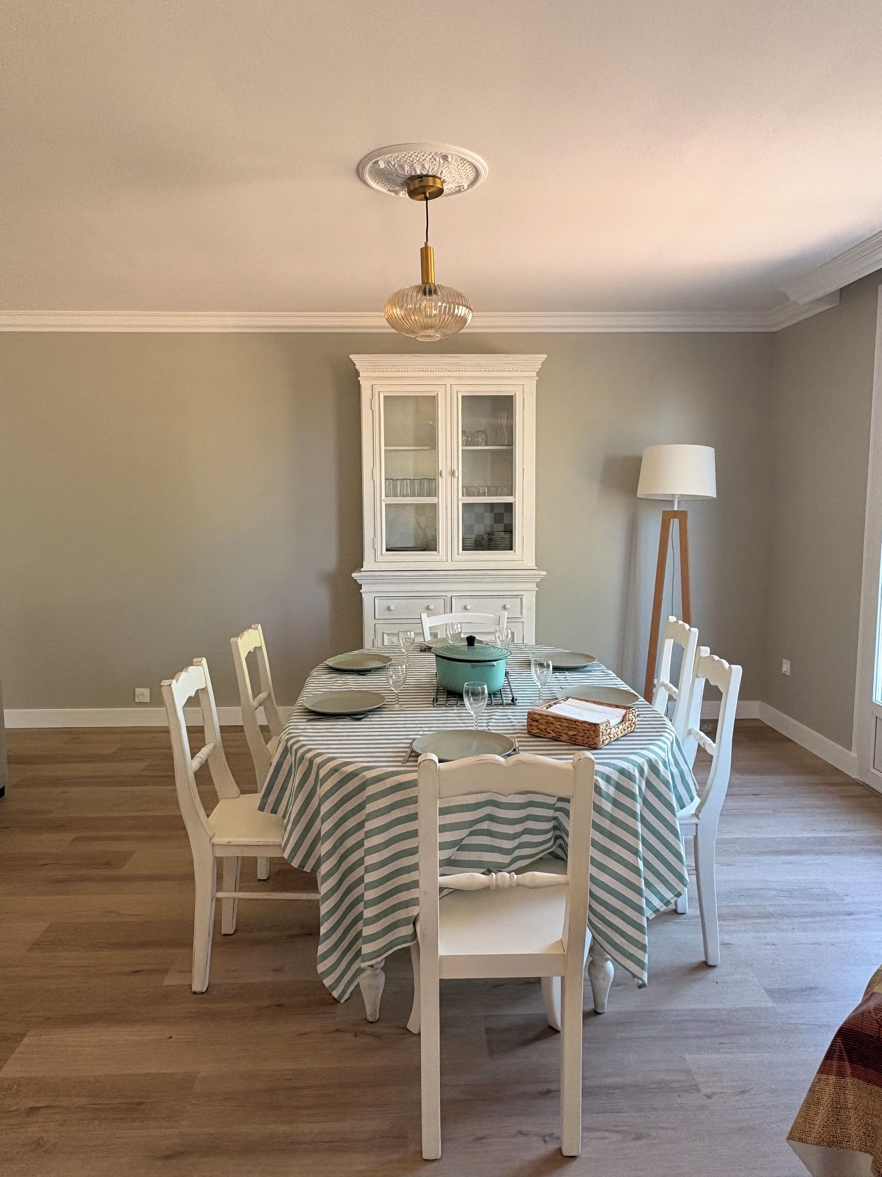 A dining room with a round table set for six, surrounded by white chairs. A white cabinet with glass doors is against the wall, and a floor lamp is beside the table.