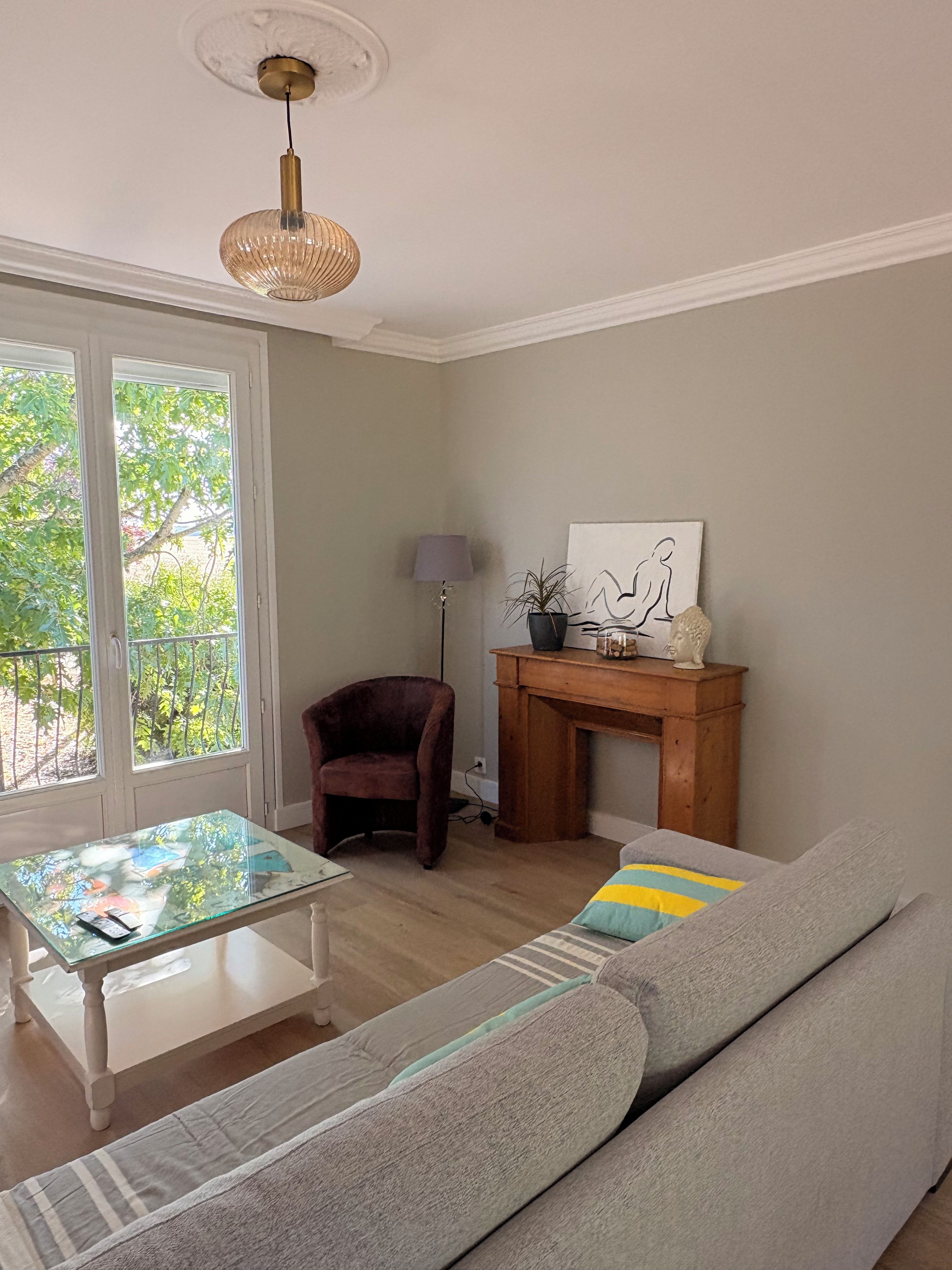 Een woonkamer met een salontafel met glazen blad, een grijze bank, een houten bijzettafeltje met een lamp en een open haard.