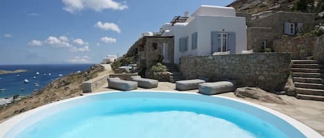 A pool area with a view of the sea, stone steps, and white buildings with blue shutters.