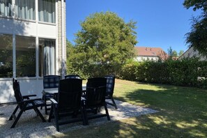 Un espace extérieur aménagé avec une table et des chaises, un bâtiment doté de grandes fenêtres et un arbre en arrière-plan.