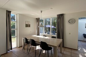 A dining area with a table and chairs, large windows, a clock, and a door leading outside. - Holiday home with 1 bedroom (Julianadorp)