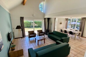 Un vaste espace de vie comprenant un canapé vert, une table basse et une table à manger avec des chaises. Un téléviseur est fixé au mur et une plante en pot est posée sur le sol.