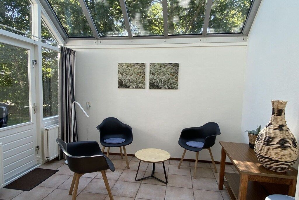 Una habitación iluminada por el sol con techo de cristal, dos cuadros en la pared, tres sillas modernas, una pequeña mesa redonda y un jarrón grande de fibra sobre una mesa de madera.