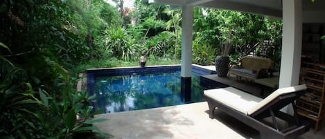 Una piscina con una silla de descanso blanca y un área de estar techada, rodeada de exuberante vegetación.