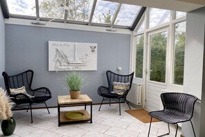 Een orangerie met dakraam, twee zwarte rieten stoelen, een houten tafel met een plant en een wandkunstwerk getiteld 'White & Free'.