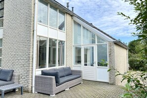 Een modern huis met een grote glazen deur, een grijze rieten bank en een metalen tafel.