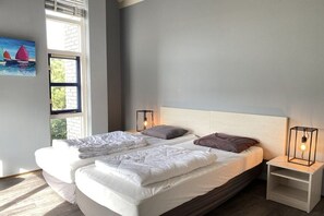 A double bed with white bedding, two bedside tables with lamps, a window with a view of sailboats, and a painting on the wall. - Holiday home with 3 bedrooms (Julianadorp aan Zee)
