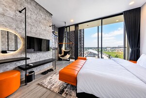 Un dormitorio moderno con una cama grande, un televisor montado en una pared texturizada, una silla colgante y vistas de la ciudad a través de grandes ventanales.