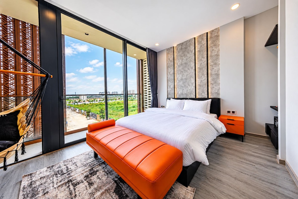 A modern bedroom with a large bed, a bench, and a view of a green landscape through floor-to-ceiling windows.