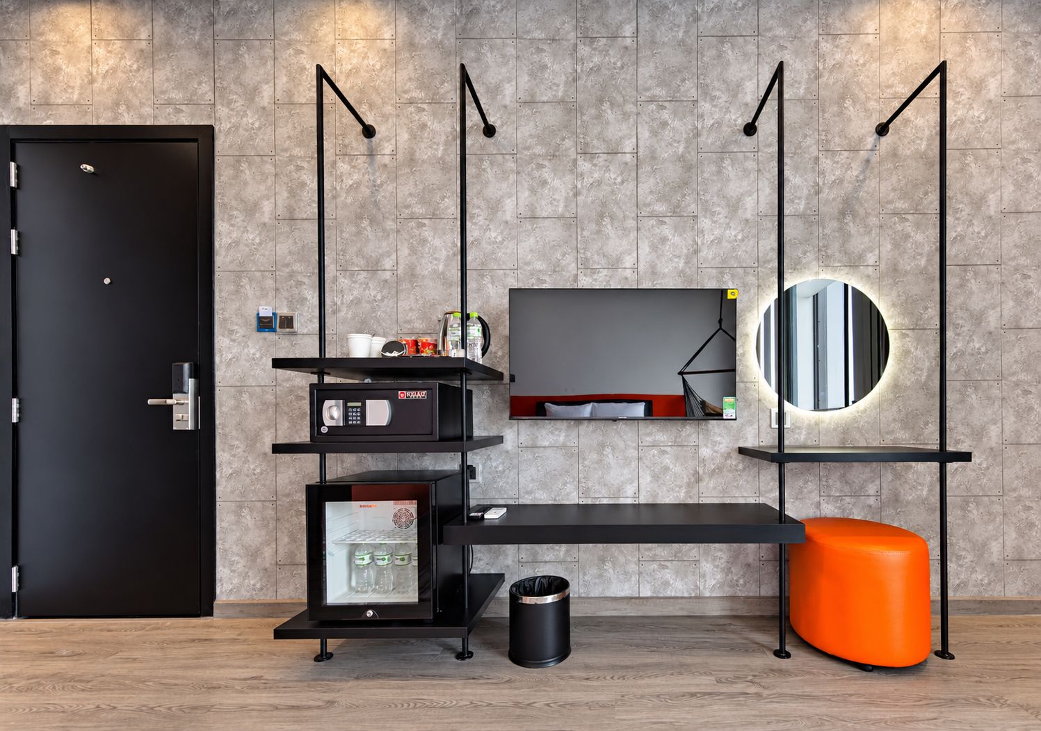 Habitación de hotel moderna con una puerta negra, un televisor de pantalla plana montado en la pared, un espejo redondo pequeño, un cubo de basura negro y un otoman de color naranja brillante.