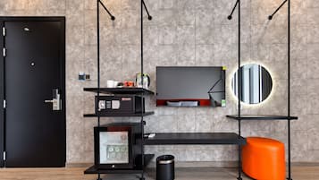 Habitación de hotel moderna con una puerta negra, un televisor de pantalla plana montado en la pared, un espejo redondo pequeño, un cubo de basura negro y un otoman de color naranja brillante.