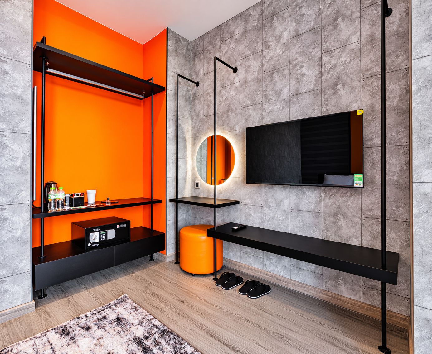 Un baño moderno con pared de azulejos grises, un estante negro con artículos de aseo, un espejo redondo con luz y un banco negro.