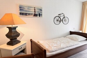 Ein Einzelbett mit Holz-Kopfteil, eine Nachttischlampe, ein kleiner Nachttisch mit Schublade, ein an der Wand befestigtes Fahrrad und ein Gemälde eines Leuchtturms.