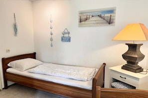 Een enkel bed met houten frame en witte beddengoed, een nachtkastje met lamp, en wanddecoraties waaronder een schilderij en een bordje.