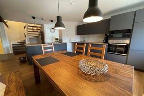 A modern kitchen with dark cabinets, a wooden dining table, and pendant lighting. - Holiday apartment with 1 bedroom (Fehmarn)