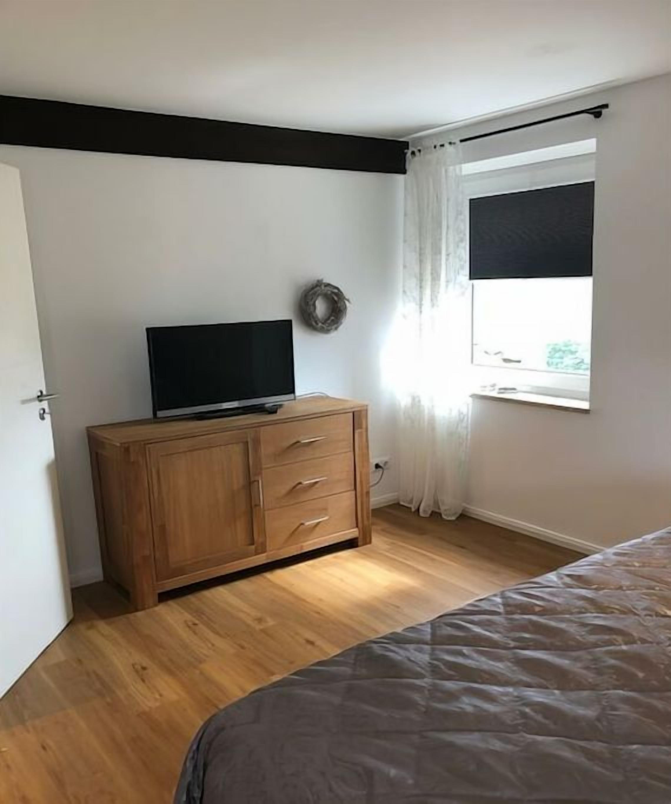 A bedroom with a bed, a wooden dresser, a television, and a window with curtains.