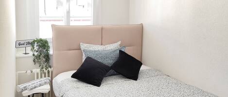 A bedroom with a bed, a bedside table, a radiator, a window, and a hanging light fixture.