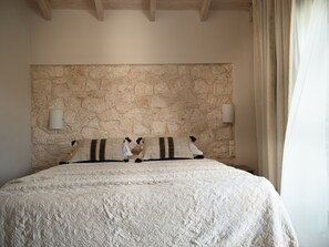 A bedroom with a stone wall headboard, a large bed with a textured white bedspread, two wall-mounted lamps, and a window with curtains. - Avesta Private Villas Niovi (Lefkada)