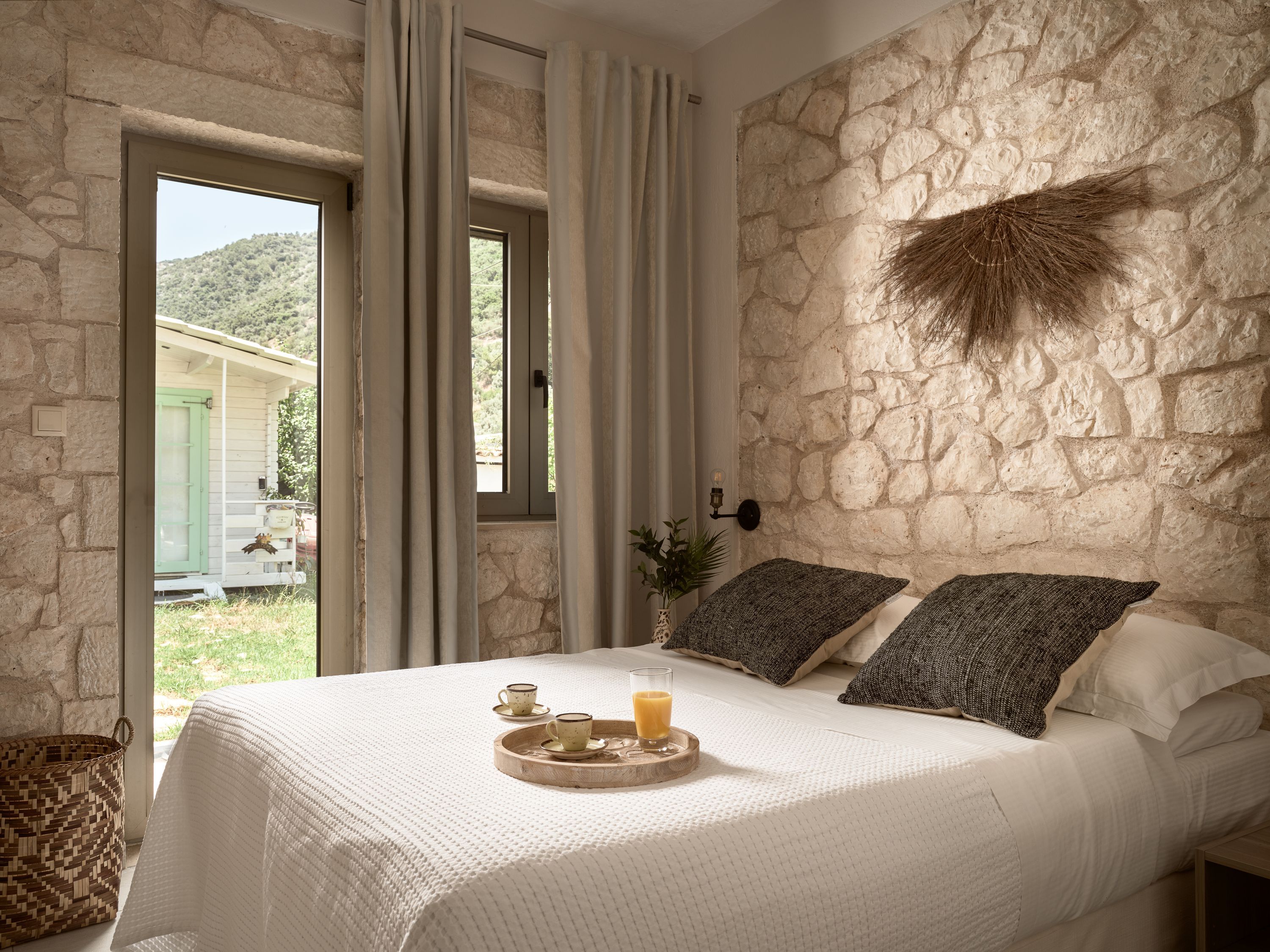 Ein Schlafzimmer mit Steinwand, einem Bett mit weißer Bettwäsche, einem Tablett mit Tassen und einem Glas Saft sowie Blick auf ein grünes Gebäude im Freien.