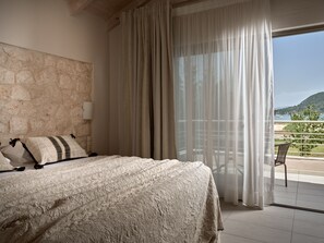 A bedroom with a stone wall, a bed with a textured blanket, a chair, and a view of a balcony with a railing and a distant landscape. - Avesta Private Villas Niovi (Lefkada)
