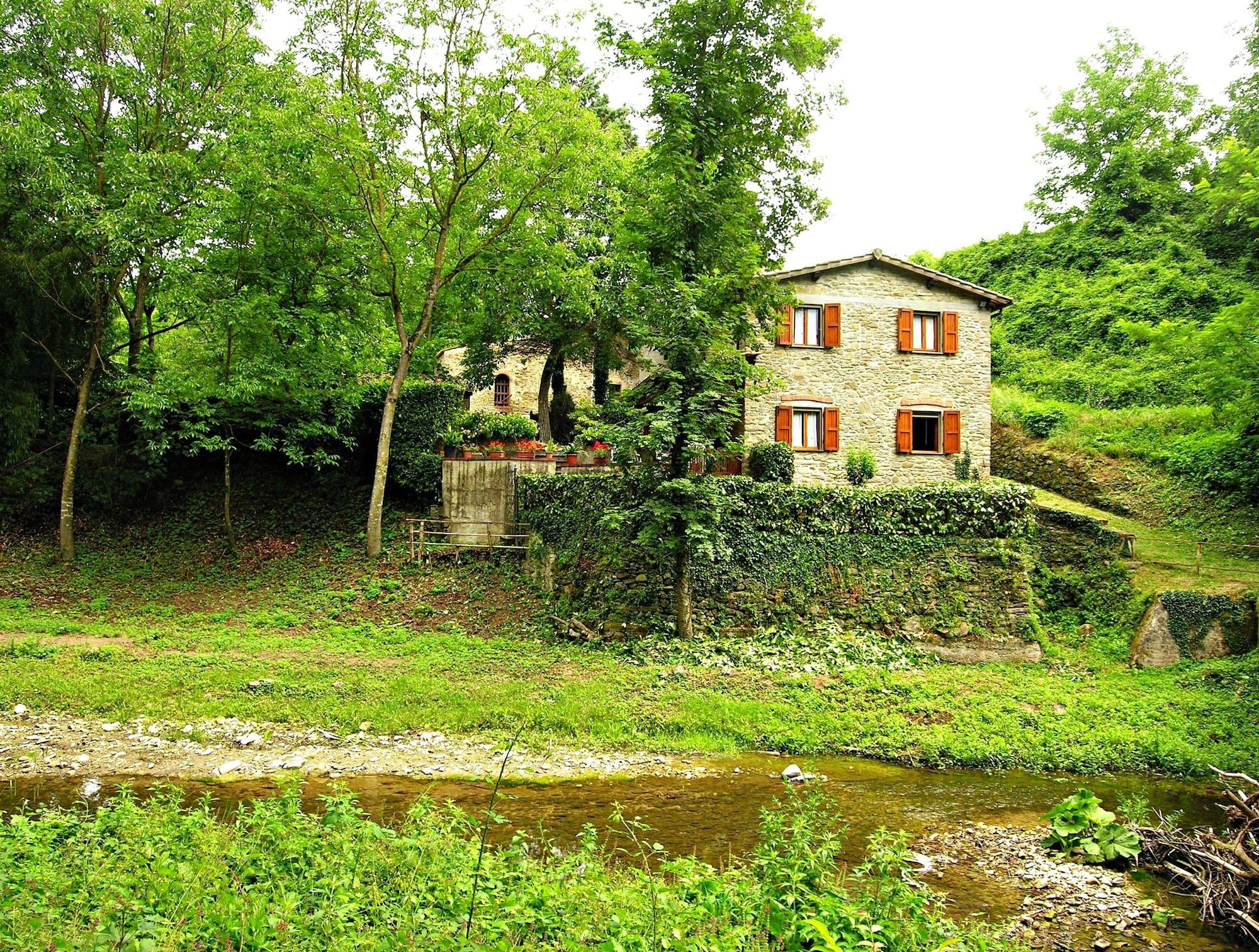 Une maison en pierre avec des volets rouges, entourée d’une végétation luxuriante et bordée d’un ruisseau.