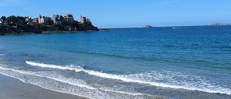 Una playa con olas, cielo despejado y edificios al fondo.