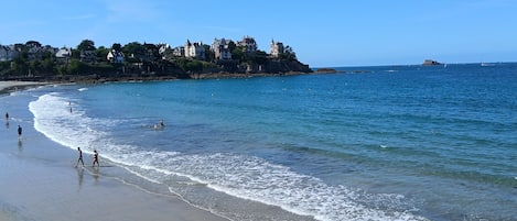 A beach with clear blue water, a sandy shore, and buildings on a cliff in the background.