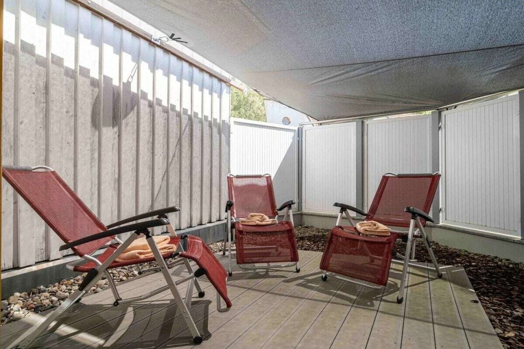 Un espace extérieur aménagé avec trois chaises rouges et une table, protégé du soleil par un auvent blanc, le tout adossé à une clôture blanche.