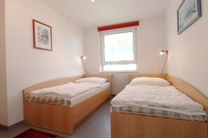 Ein Zimmer mit zwei Holzbetten, einem Fenster mit Jalousien, einem gerahmten Bild an der Wand und einem roten Akzent an der Decke.