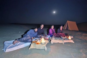 A group of people sitting on the ground at night with a tent and a moon in the background. - The Chirag Desert Camp (Jaisalmer)