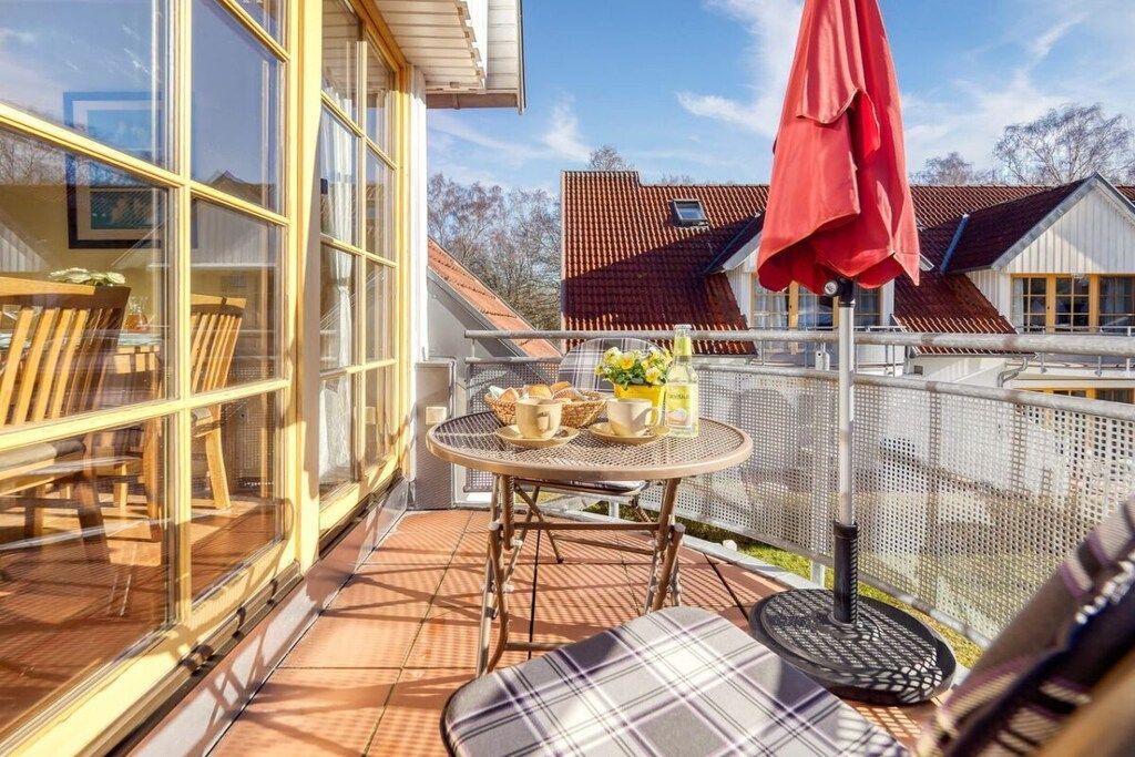 Un balcon ensoleillé avec une table dressée pour deux personnes, un parasol rouge et une vue sur les maisons voisines.