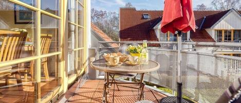 Un balcon ensoleillé avec une table dressée pour deux personnes, un parasol rouge et une vue sur les maisons voisines.