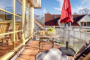 Un balcon ensoleillé avec une table dressée pour deux personnes, un parasol rouge et une vue sur les maisons voisines.
